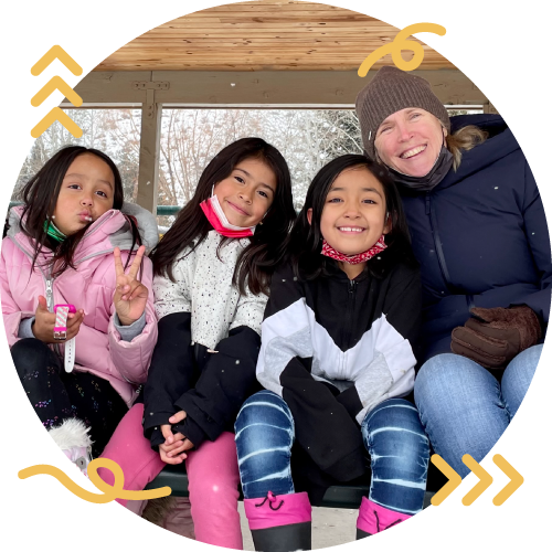 Four young girls and an adult woman sitting together outdoors in cold weather, smiling and making peace signs, with a wooden shelter and trees in the background.