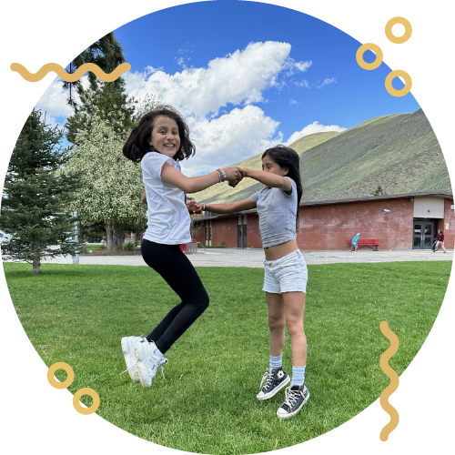 Two young girls are playing and holding hands in a green park with a building and hills in the background on a sunny day.