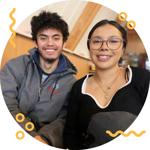 Two young adults sitting together at a table, smiling, in an indoor setting with wood-paneled walls.