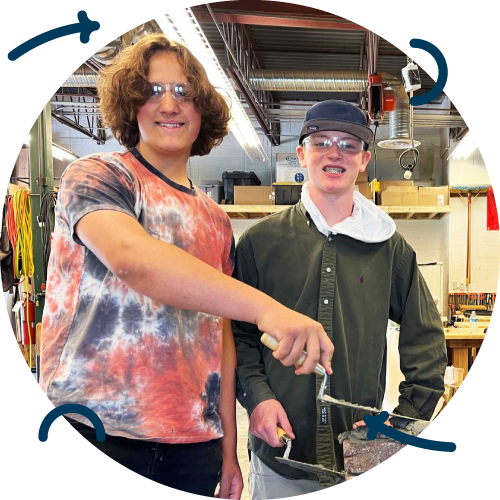 Two smiling people, one with curly hair and sunglasses wearing a tie-dye shirt, and the other younger with glasses, a cap, and a green jacket, standing in a workshop with tools, shelves, and a ceiling with exposed pipes.