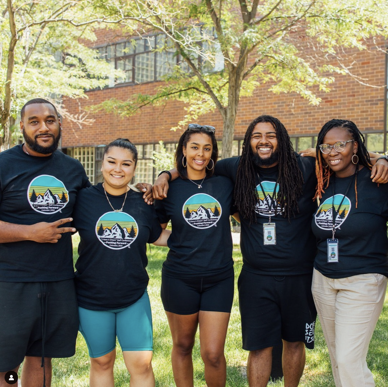Group of five young adults standing outdoors with trees and a brick building in the background. They are smiling, wearing matching black T-shirts with a mountain graphic, and have their arms around each other.