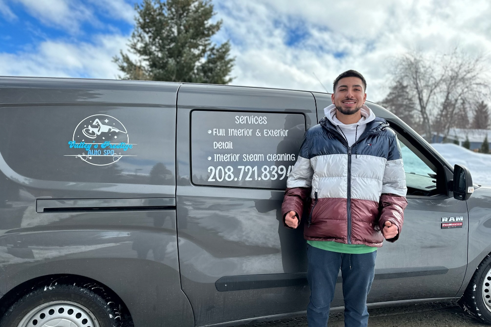 A smiling man standing next to a gray auto detailing van outdoors in winter. The van has a logo with a moon and stars, and text listing services and a phone number.