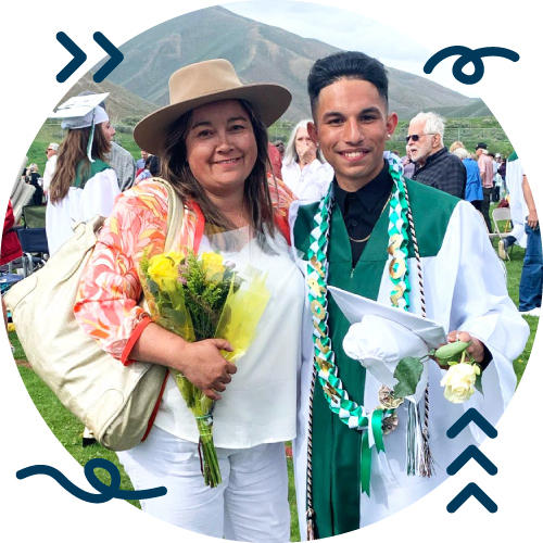 A woman in a tan hat and floral cardigan holding a bouquet, standing next to a young man in graduation attire with a cap and gown, at an outdoor graduation ceremony with mountains in the background.