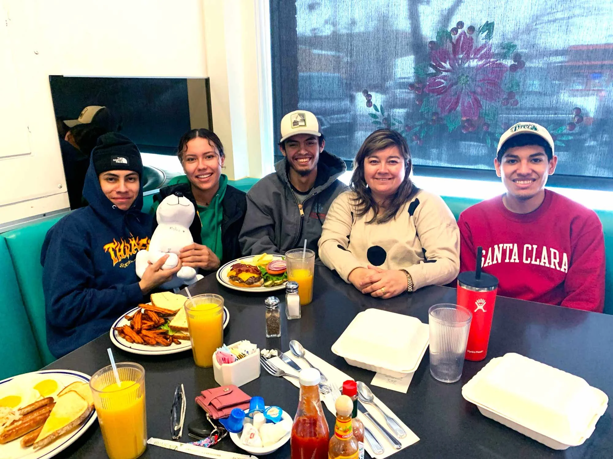 Group of five people sitting at a restaurant table with breakfast items, including two plates of toast and sweet potato fries, and drinks like orange juice. They are smiling, and the person on the left is holding a plush toy, in front of a window with a flower design.