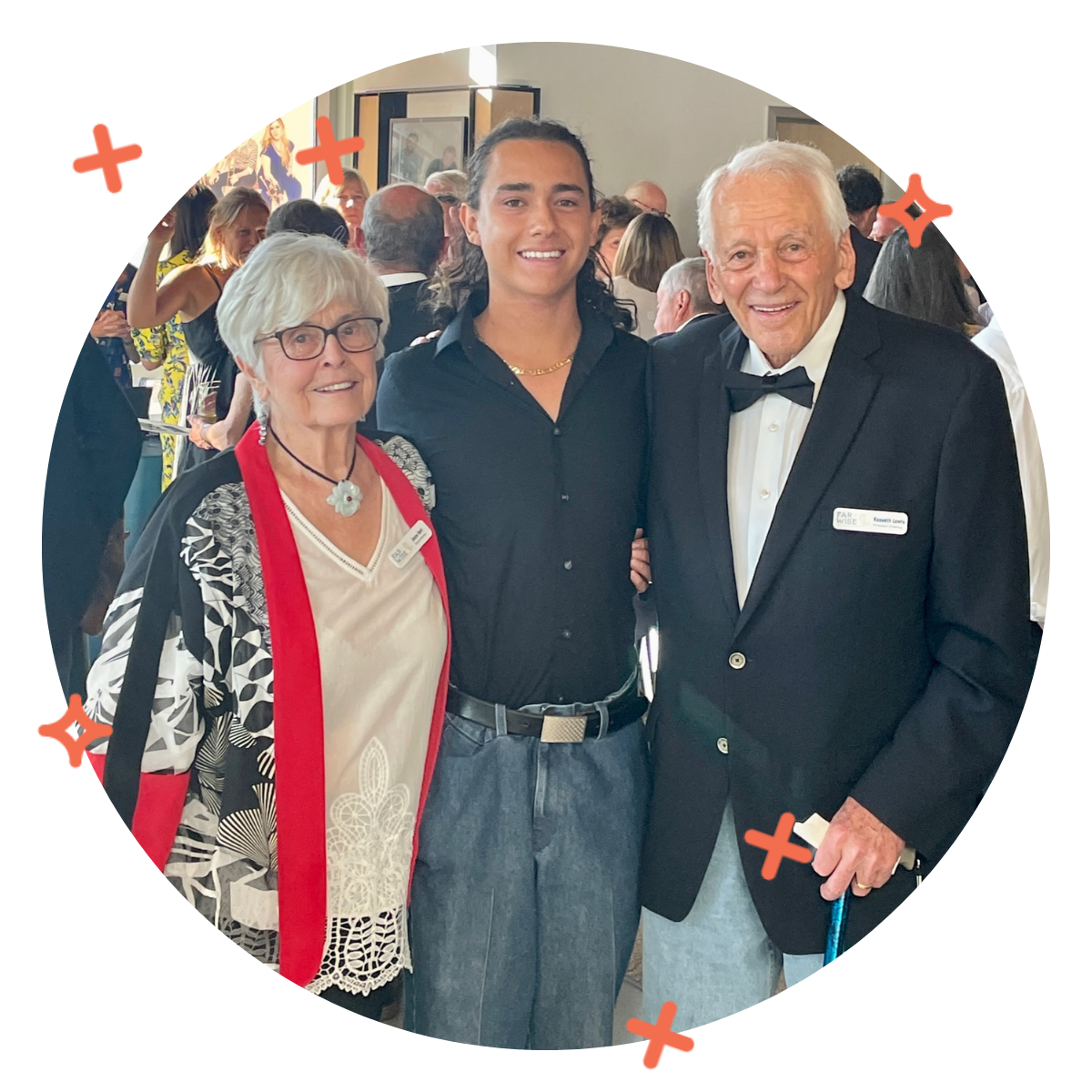 Three people, an elderly woman, a young man, and an elderly man, dressed in formal attire, smiling at a social event with a room full of other guests in the background.