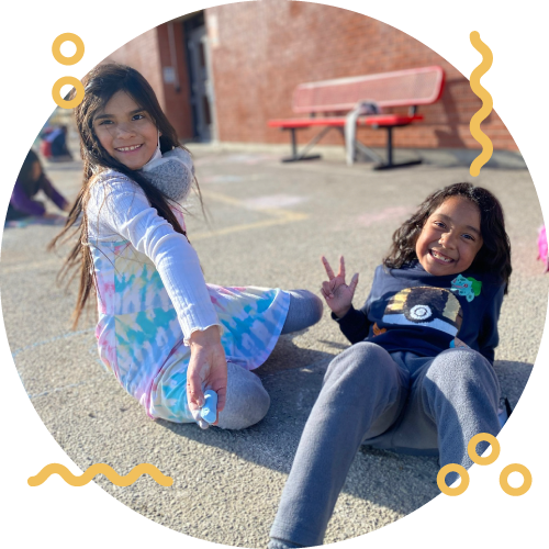 Two young girls sitting on the ground outdoors, smiling and making peace signs. One girl is wearing a pastel dress and the other a hoodie and sweatpants.