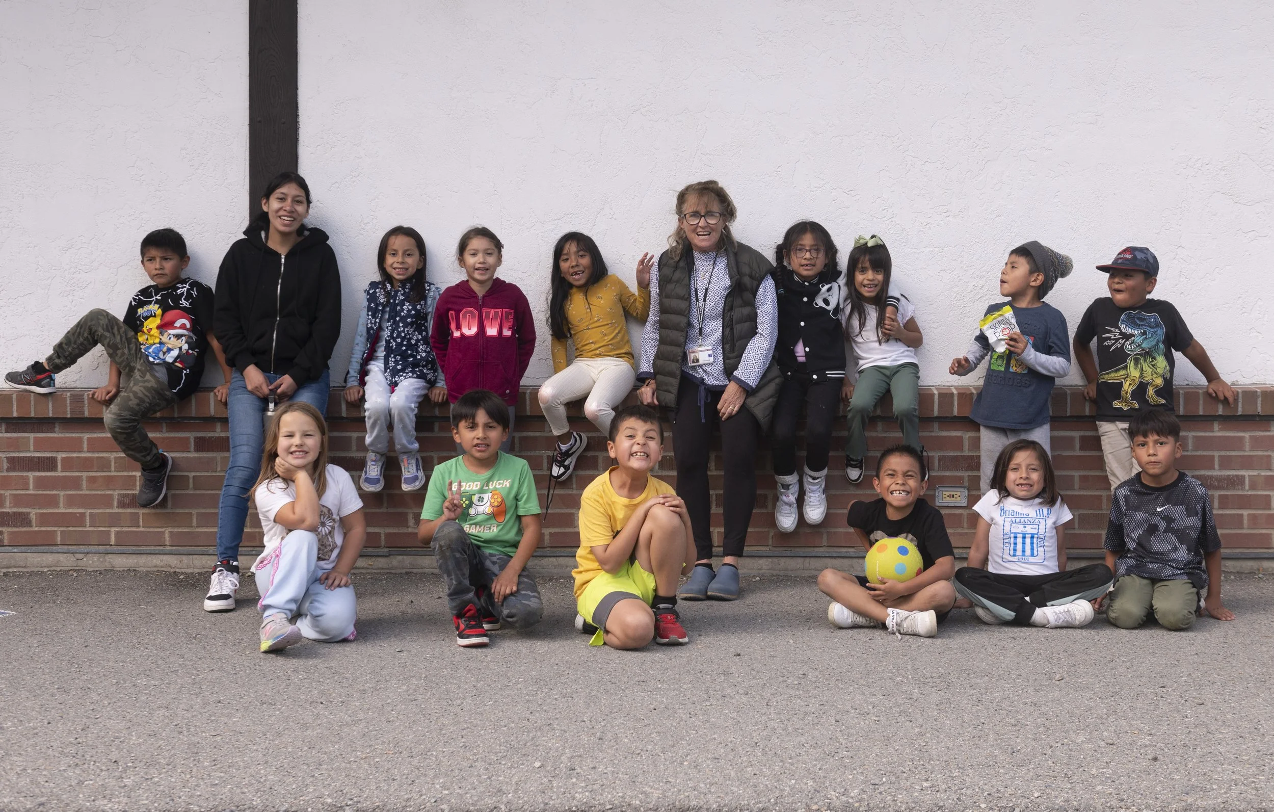 Group of children and an adult woman sitting and kneeling outside in front of a white wall, smiling and posing for the photo.
