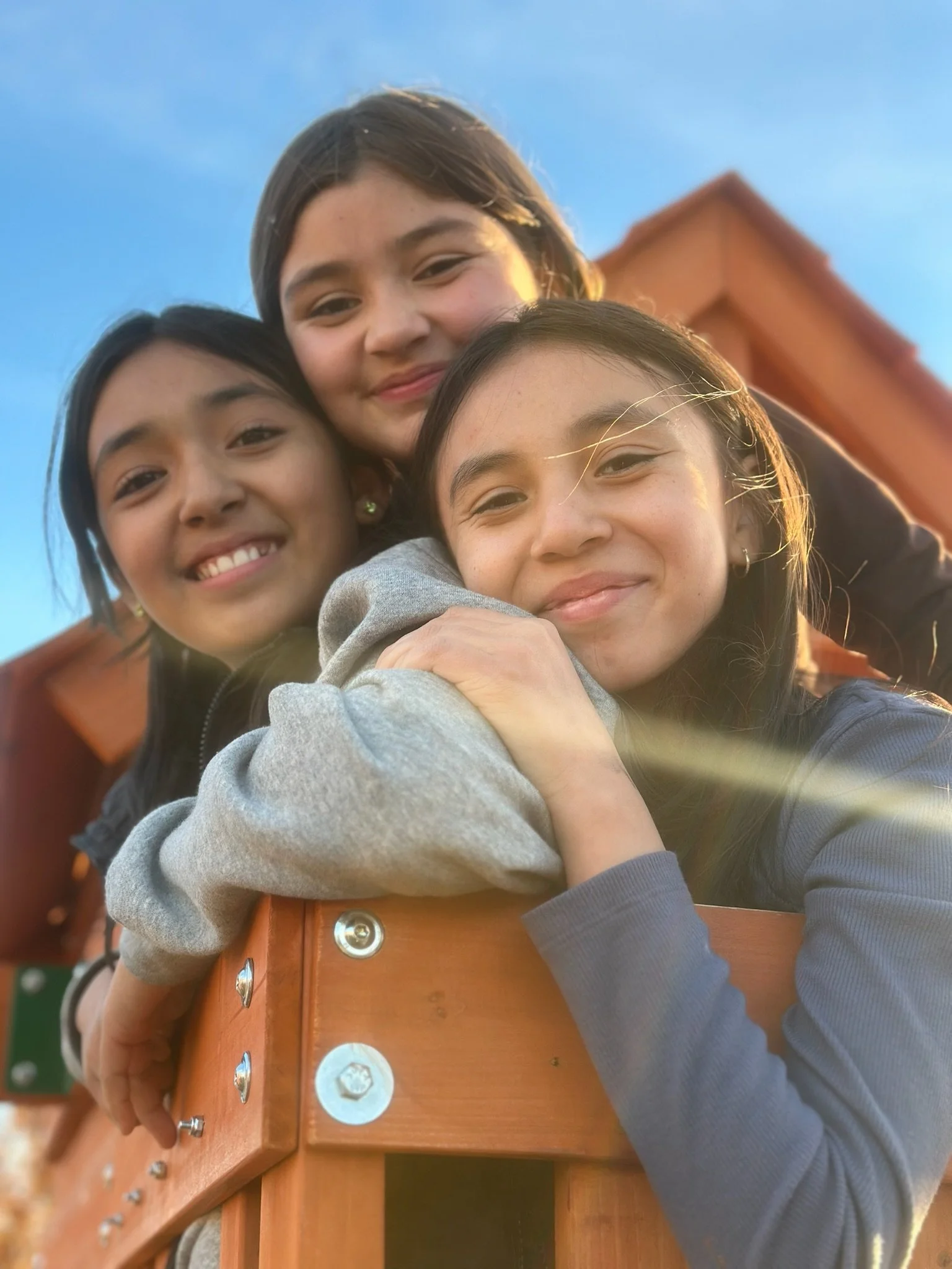Three girls happily hugging and smiling on a wooden playground structure under a clear blue sky.
