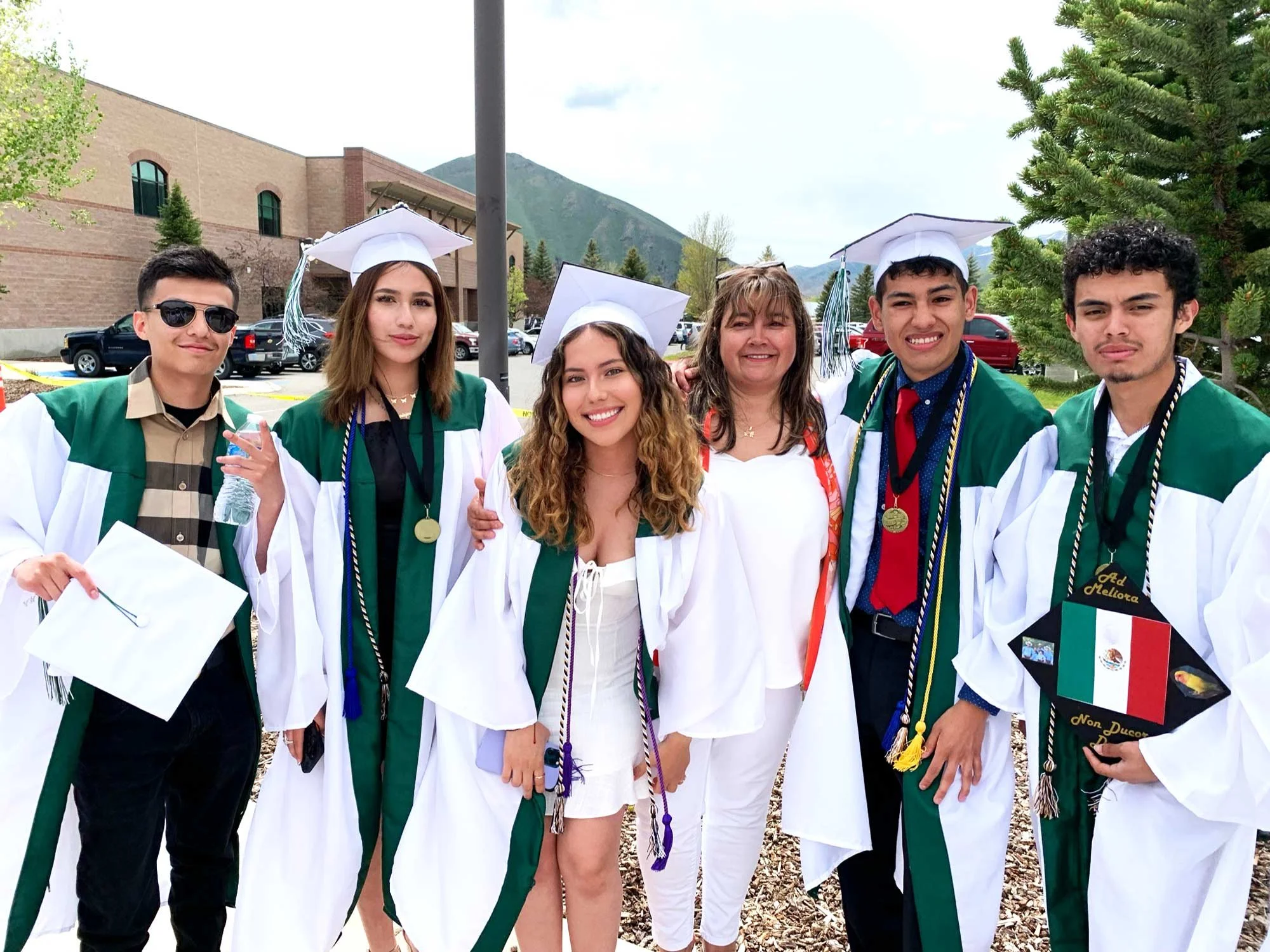 Group of six young adults celebrating graduation outdoors, wearing green and white caps and gowns, with friends and family, and mountain scenery in the background.