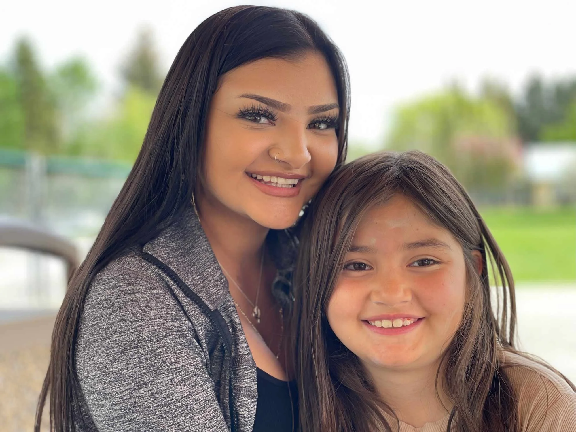 A woman with long dark hair and a septum piercing, smiling, standing next to a girl with long brown hair, both smiling, outdoors with greenery in the background.