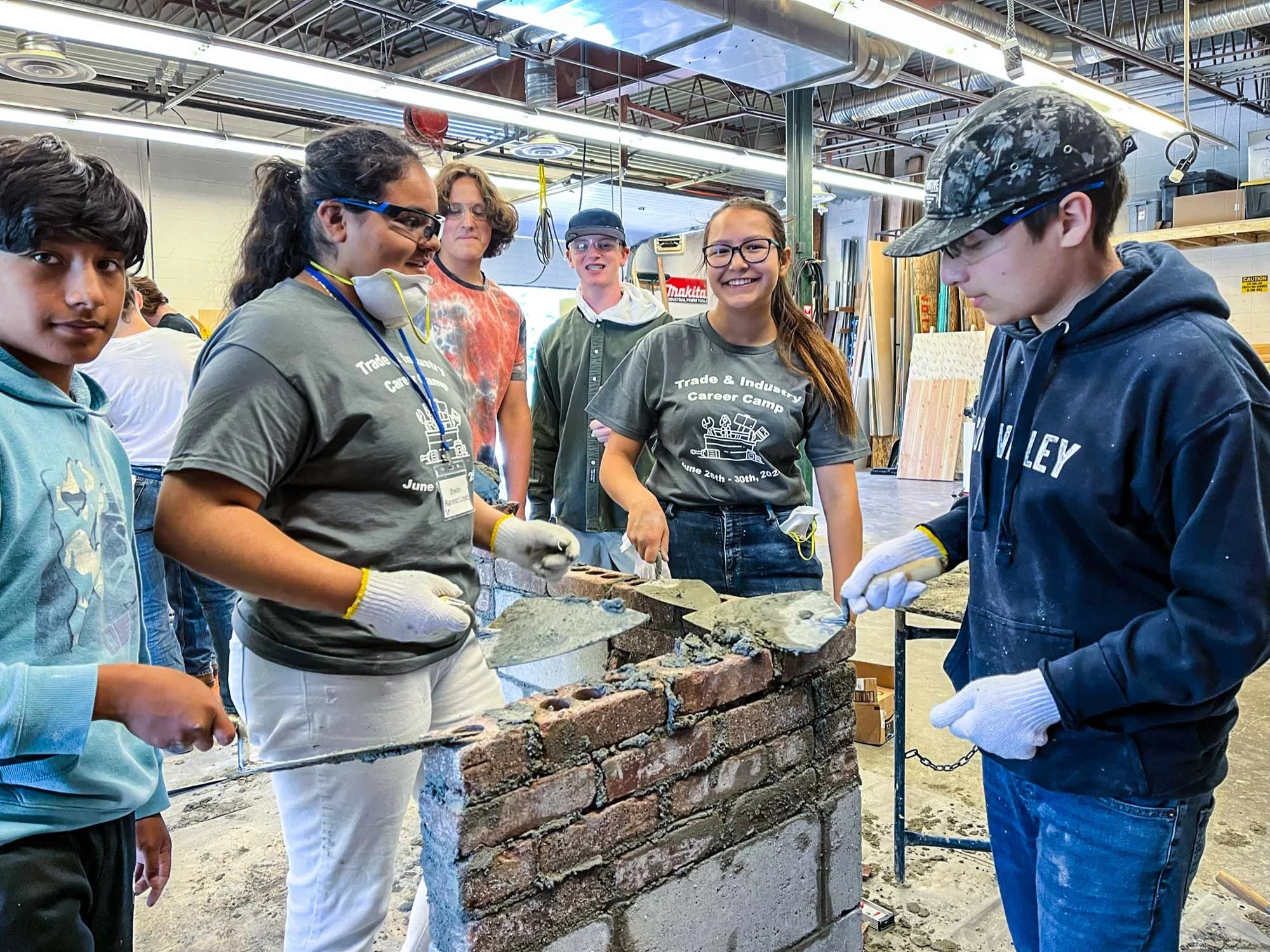 Group of students working on a brick wall project in a woodworking workshop, some wearing gloves and safety gear, others observing or smiling.