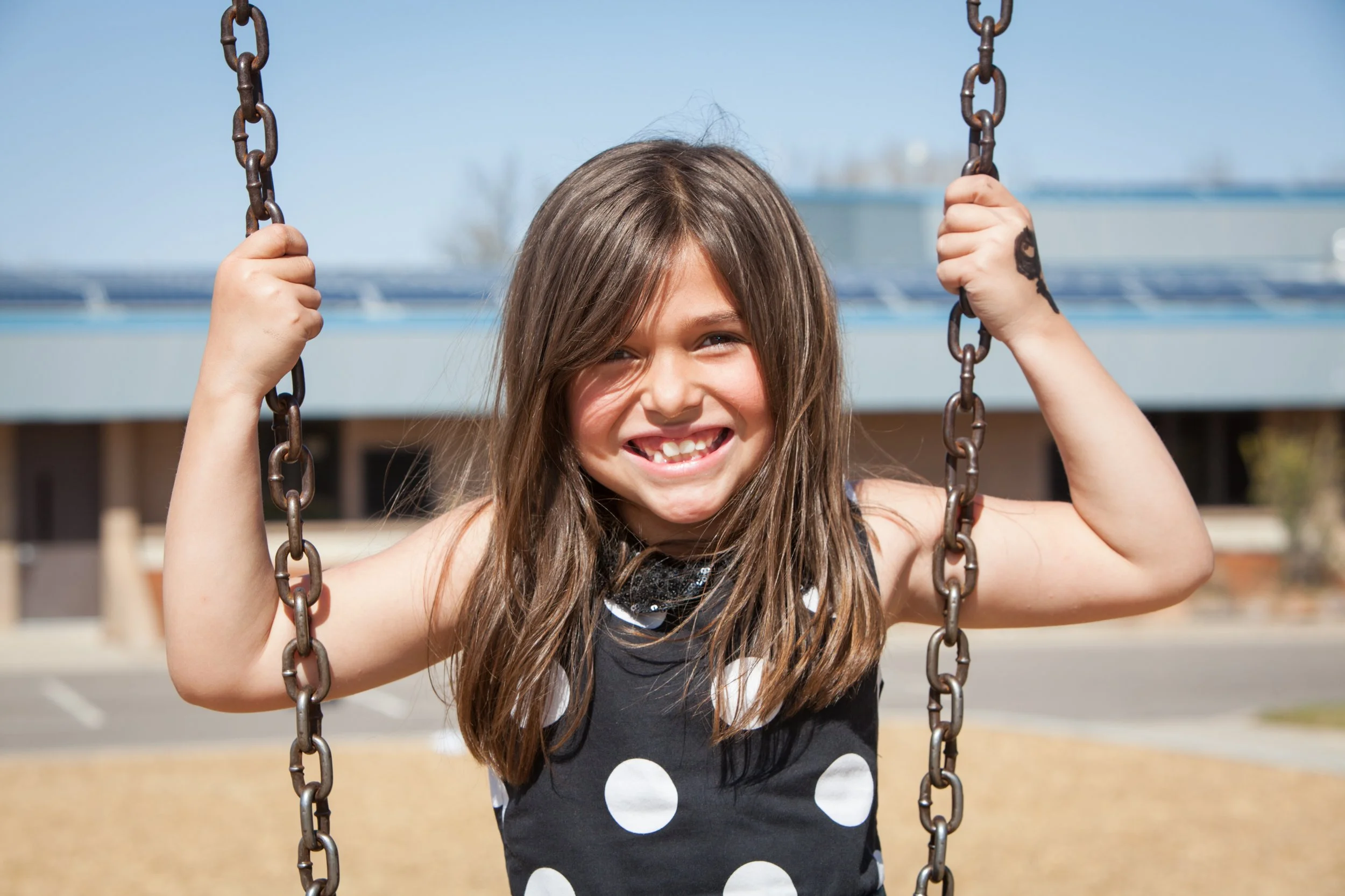A young girl with long brown hair smiling and holding onto a swing set's chains at a park.