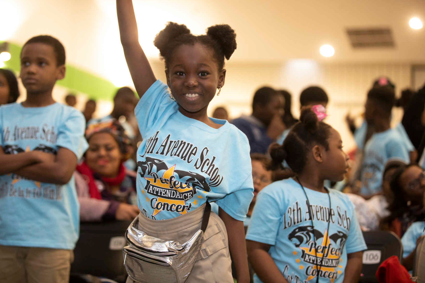 A young girl with dark hair styled in two puffs, smiling and raising her hand. She is wearing a light blue T-shirt with '13th Avenue School' and other event text, beige pants, and a metallic purse. The background shows other children and adults attending the same event, some wearing similar T-shirts.