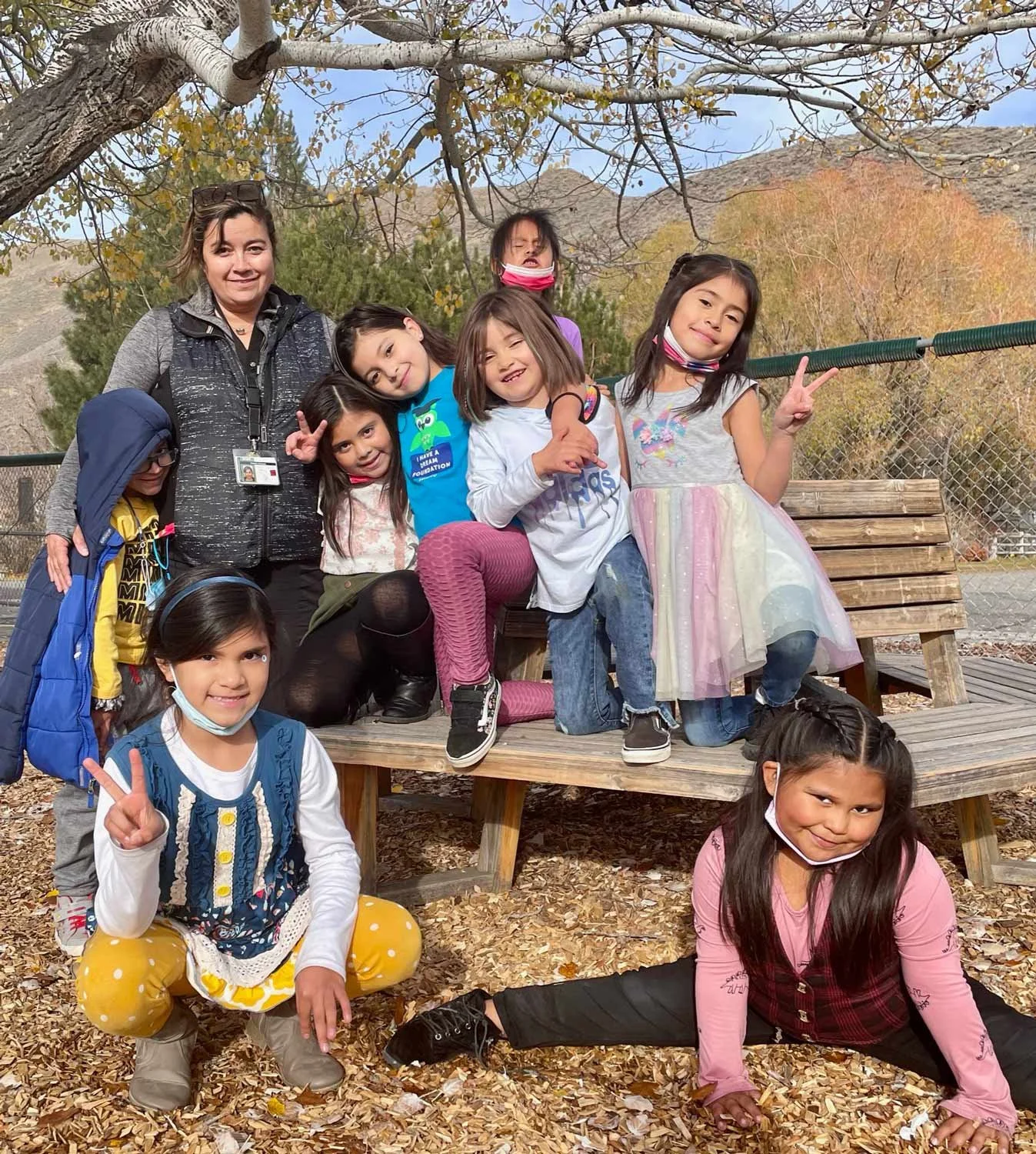 Group of children and one adult woman outdoors, sitting and posing for a photo on a wooden bench and ground, with autumn trees and mountains in the background.