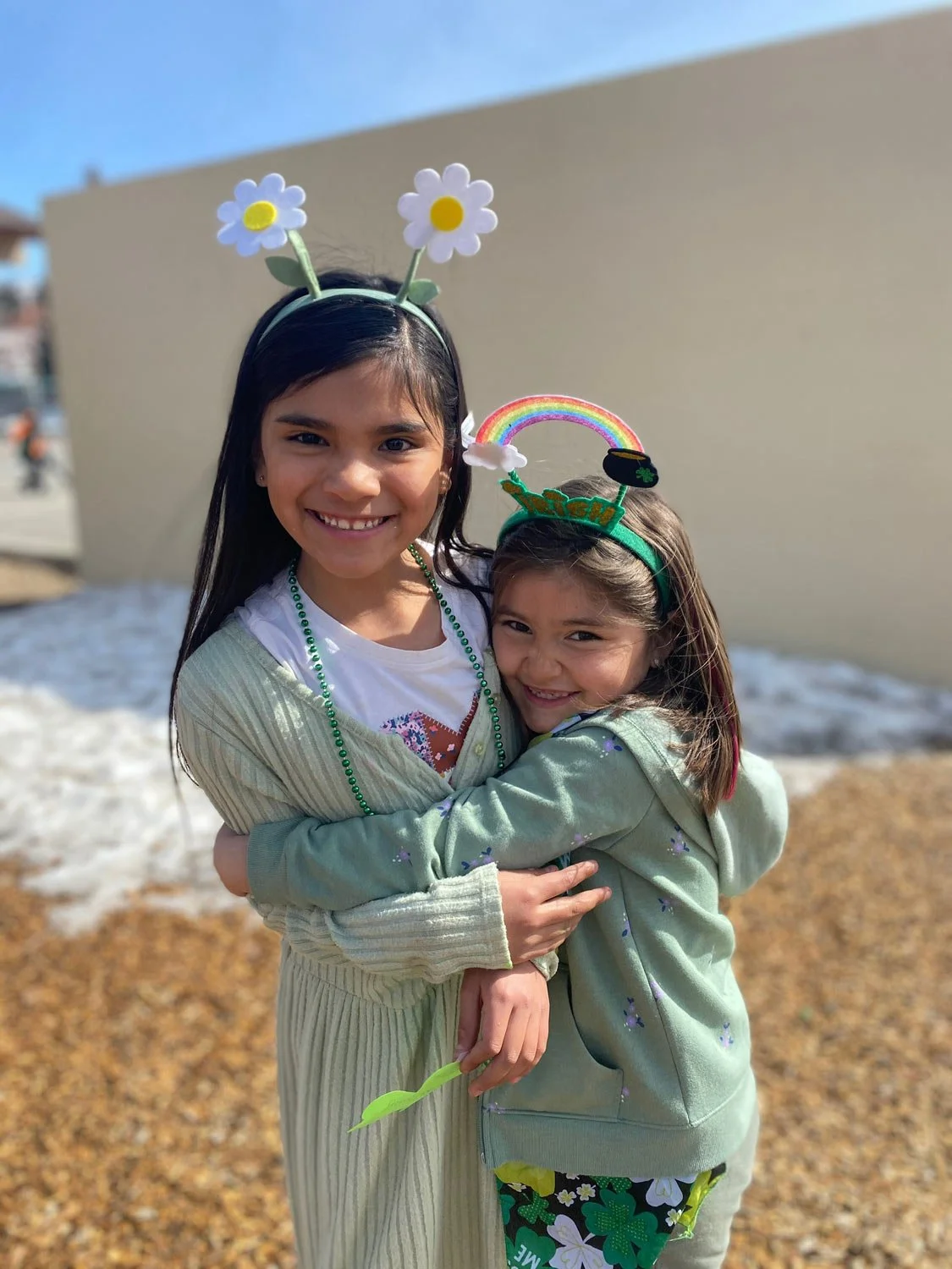 Two young girls hugging outdoors on a sunny day, wearing festive headbands with rainbow, flowers, shamrocks, and rainbows, celebrating St. Patrick's Day.
