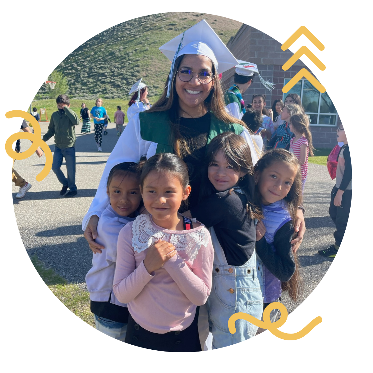 A woman in graduation cap and gown with four young girls, outdoors at a graduation event, with other children and adults in the background.