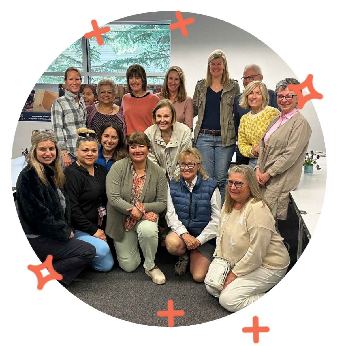 A diverse group of women and two men gathered together indoors for a group photo, smiling and posing for the camera. Some are standing, while others are kneeling or sitting in front. There is a large window in the background with greenery outside.