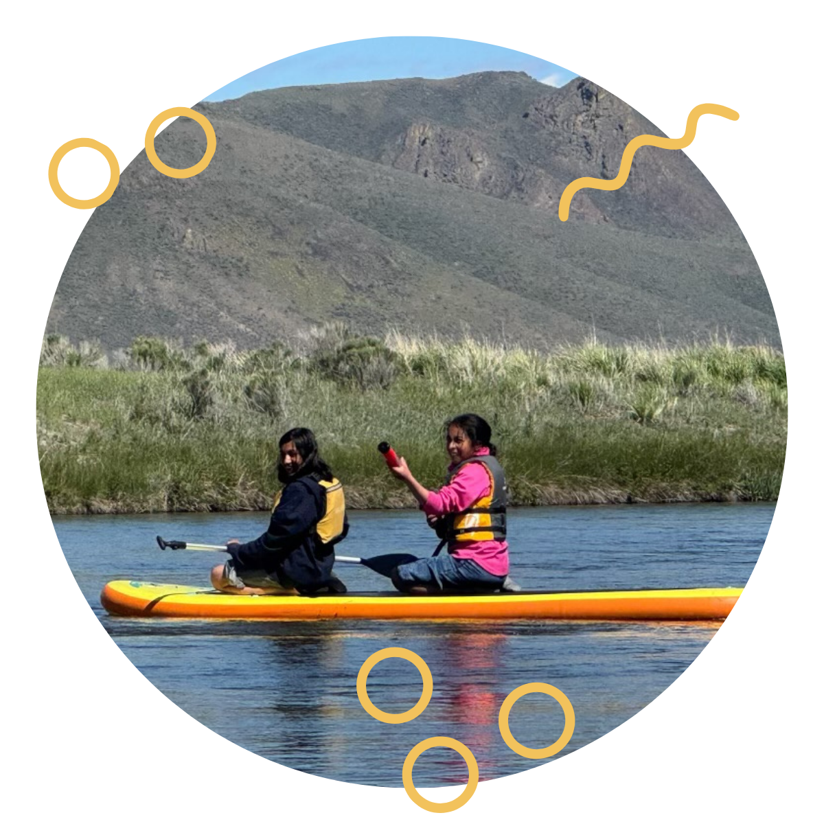 Two young women kayaking on a lake with mountains and grassy landscape in the background.