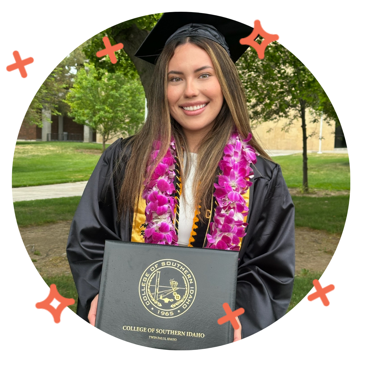 A young woman in a black graduation cap and gown holding a diploma from the College of Southern Idaho in Twin Falls, Idaho, with a pink lei around her neck, standing outdoors with trees and a building in the background.