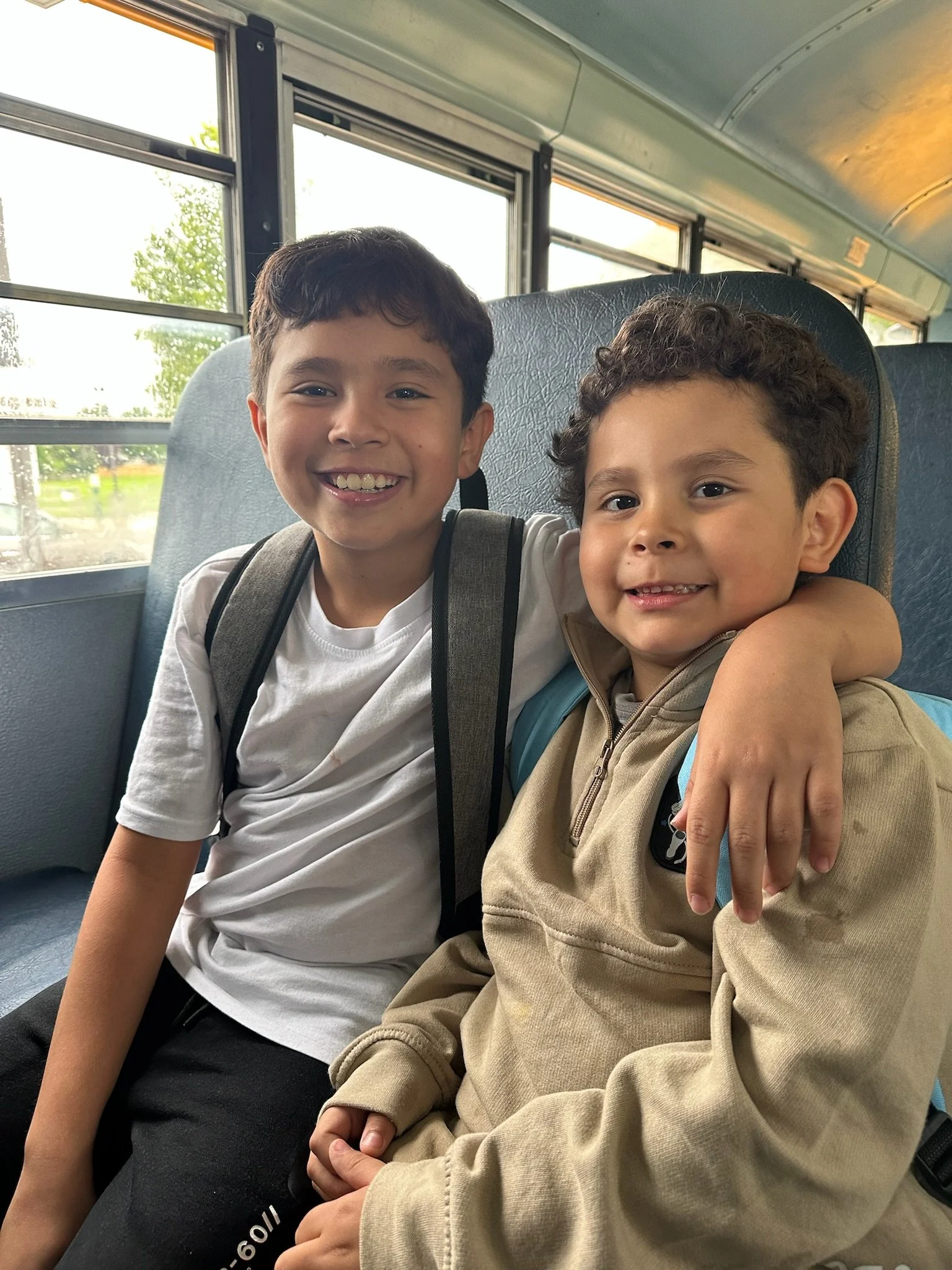 Two young boys sitting close together on a school bus, smiling at the camera, with one boy's arm around the other's shoulder.