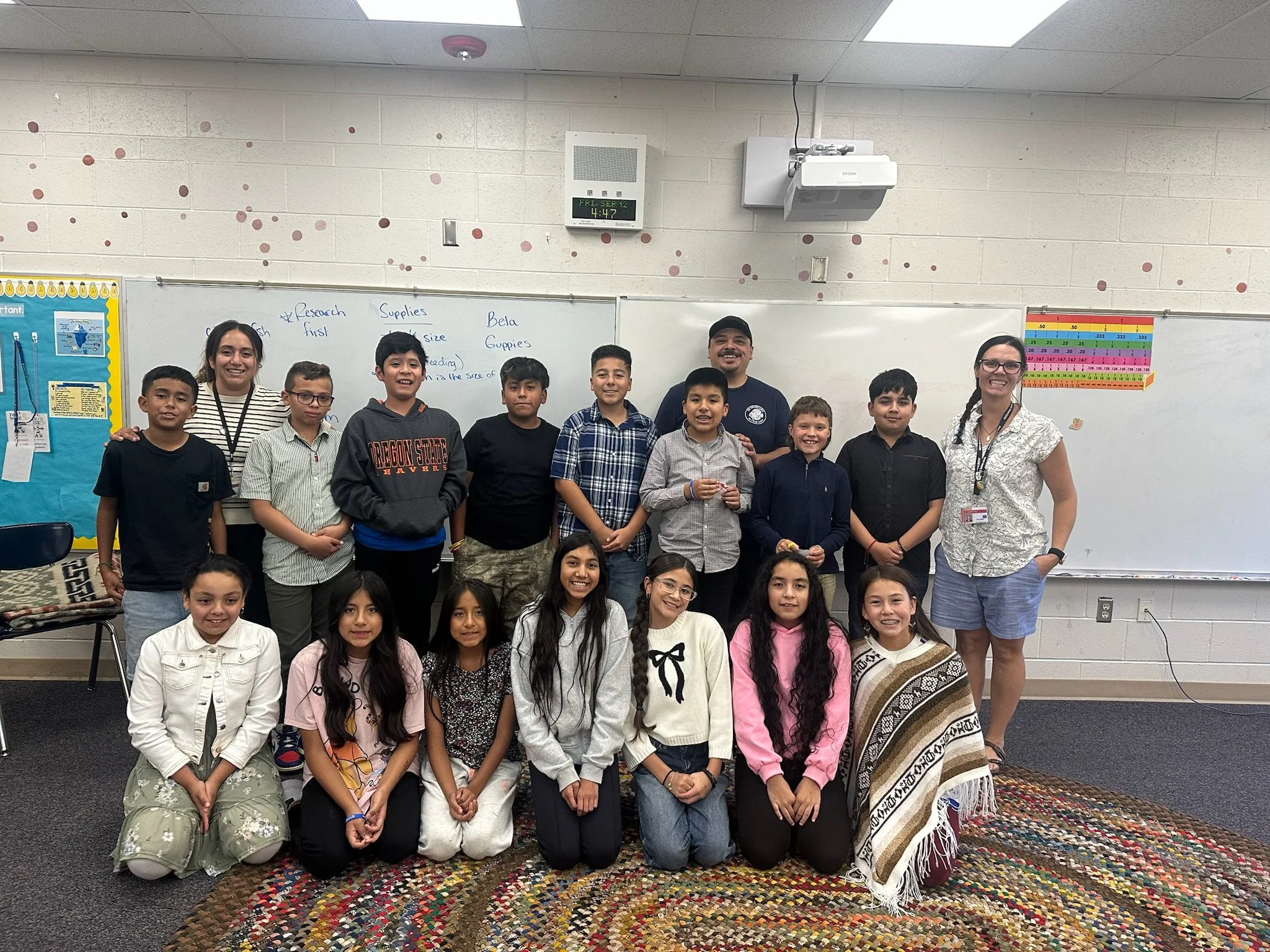 A group of young students and two adults, likely teachers, posing for a classroom photo in front of a whiteboard with notes about research and supplies. The children are sitting and standing, smiling. The classroom has educational posters, a colorful rug, and a whiteboard with writing.