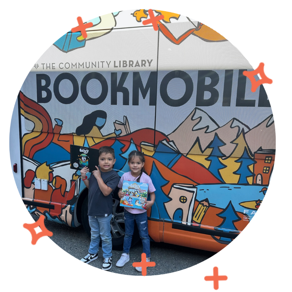 Two children standing in front of a colorful mobile library truck with mountain and forest illustrations, holding books and smiling.