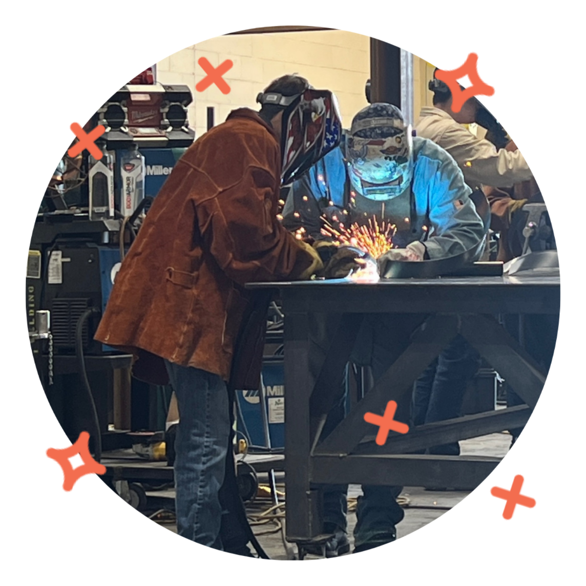 Two workers welding sparks fly while working at a table in a workshop, both wearing protective gear including helmets and gloves.