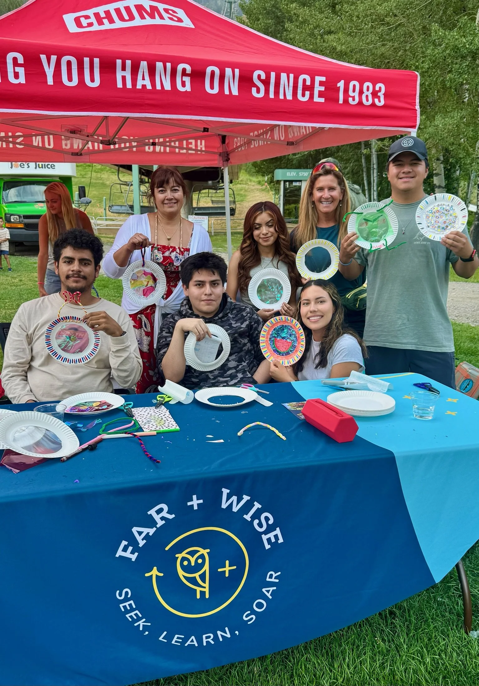 Group of people at an outdoor craft event under a red tent that reads "CHUMS" and "Thanks for hanging on since 1983." They are displaying decorated paper plates and sitting behind a table covered with a blue tablecloth that says "Fear + Wise - Seek, Learn, Soar." The group consists of children and adults, smiling and showing their craft projects.