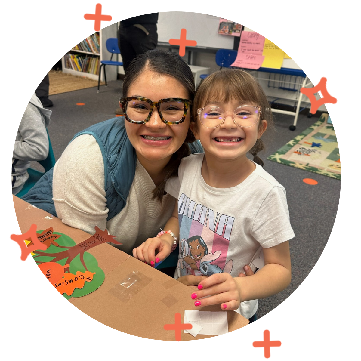 A woman and a young girl smiling at the camera inside a classroom, with bookshelves and colorful posters in the background.