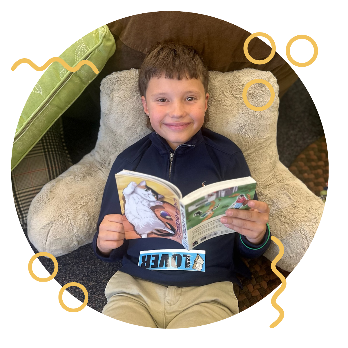 Young boy sitting in a cozy chair, smiling, holding a colorful children's book with a cartoon cat on the cover.
