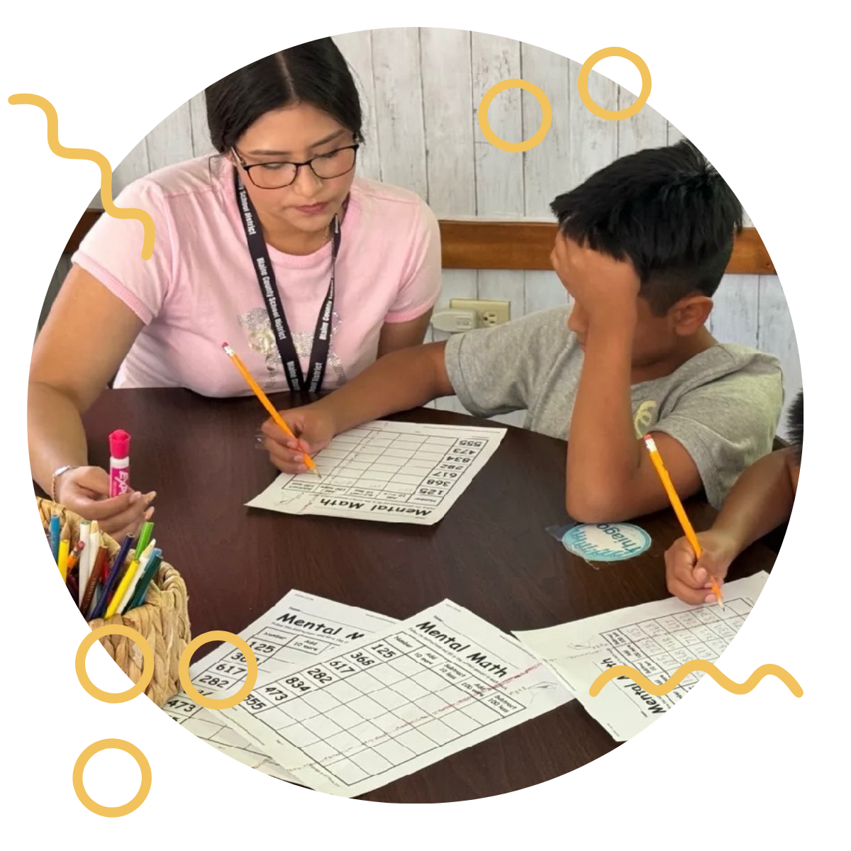 A teacher working with a young boy on a mental math activity at a classroom table, with educational papers and colorful pens nearby.