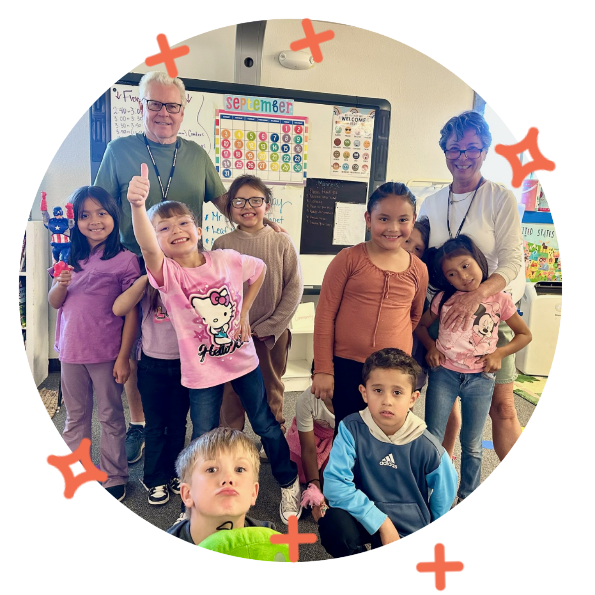 A group of children and two adults in a classroom, smiling and posing for a photo, with a colorful calendar and educational posters in the background.