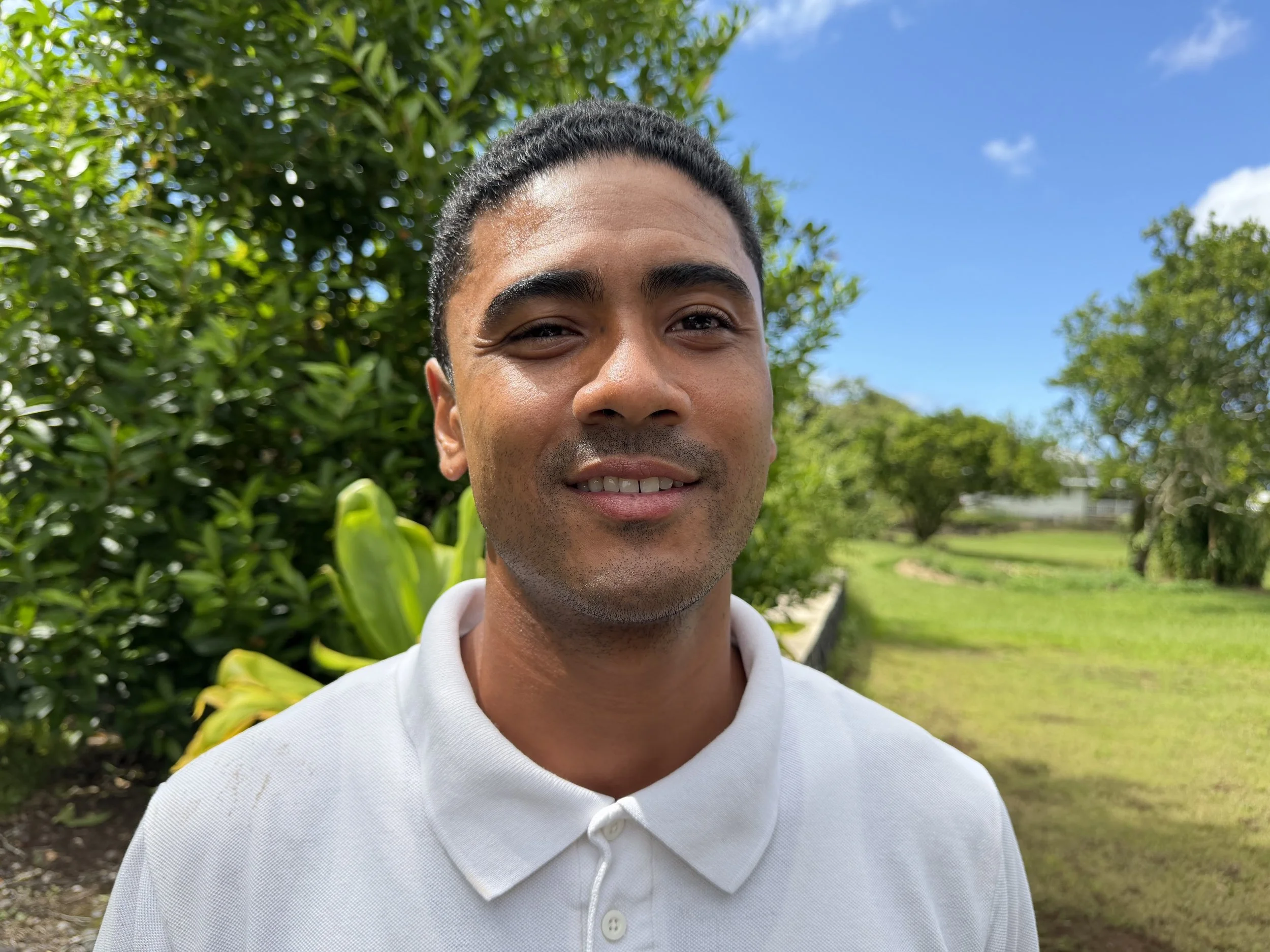 A young man smiling outdoors with green trees and a blue sky in the background. He has short dark hair, tan skin, and is wearing a white polo shirt.
