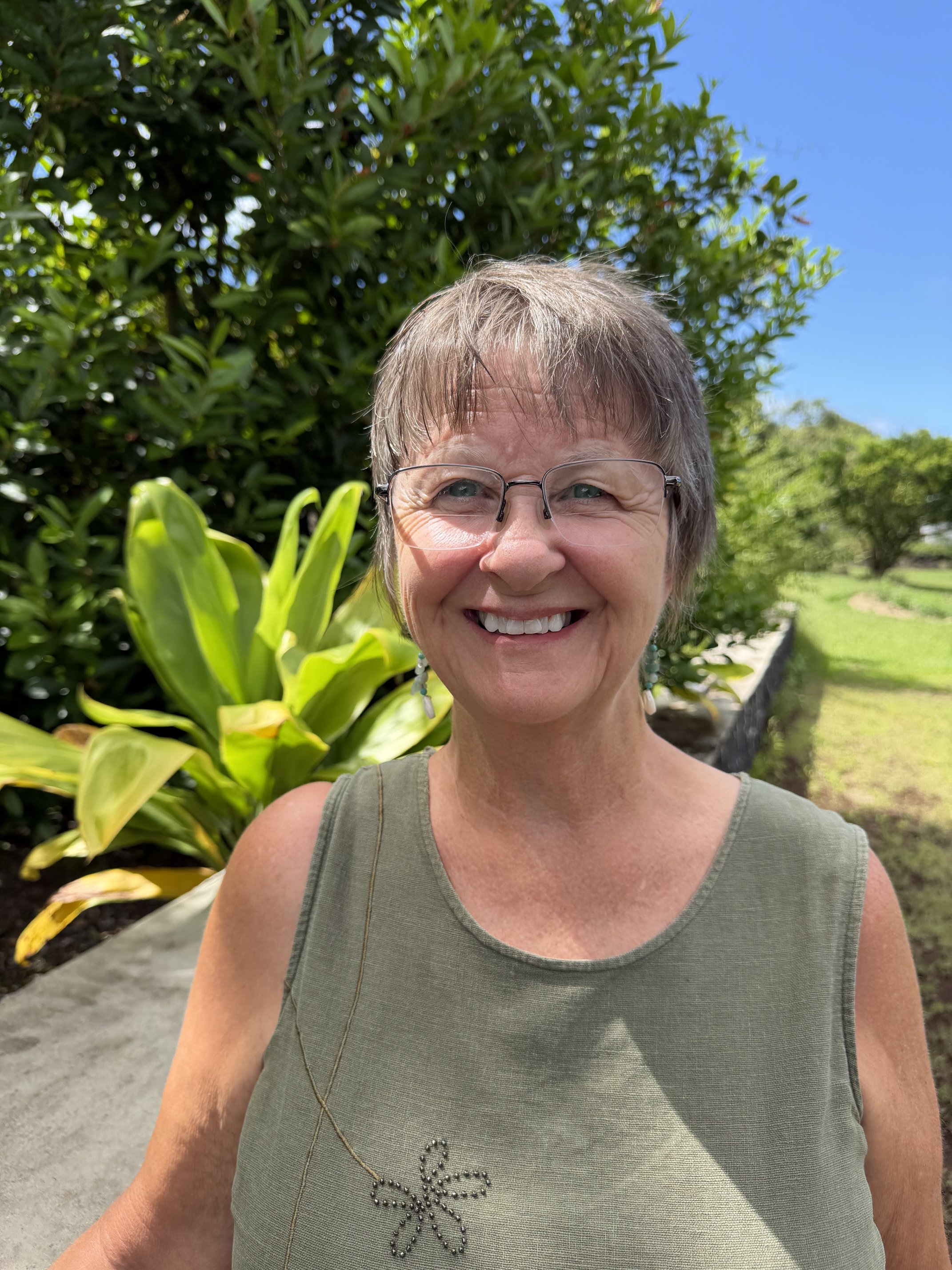 A smiling woman with short gray hair and glasses taking a selfie outdoors, with green foliage and a blue sky in the background.