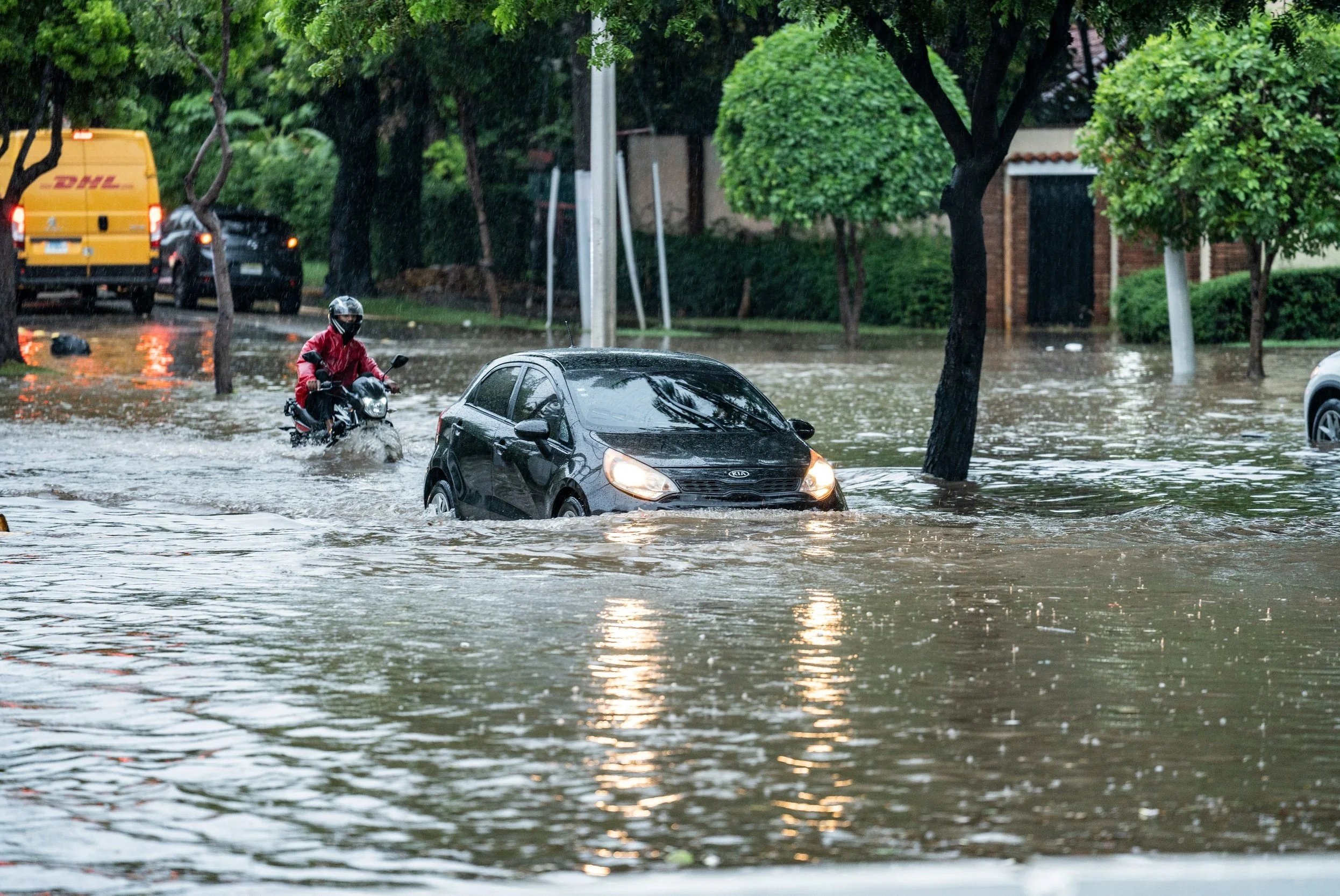 A street flooded with water, with a man riding a motorcycle and a car partially submerged. In the background, a yellow DHL delivery van, parked cars, green trees, and houses are visible.