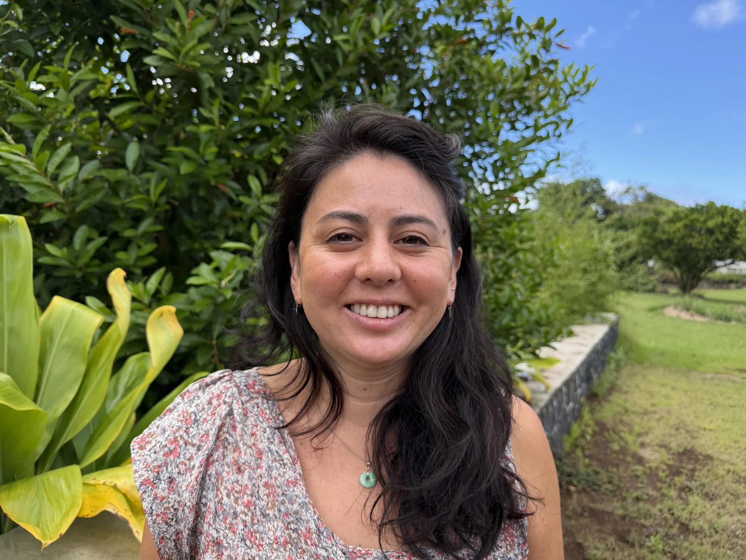 A woman with dark hair smiling outdoors, background of green foliage, blue sky, and flowering plants.
