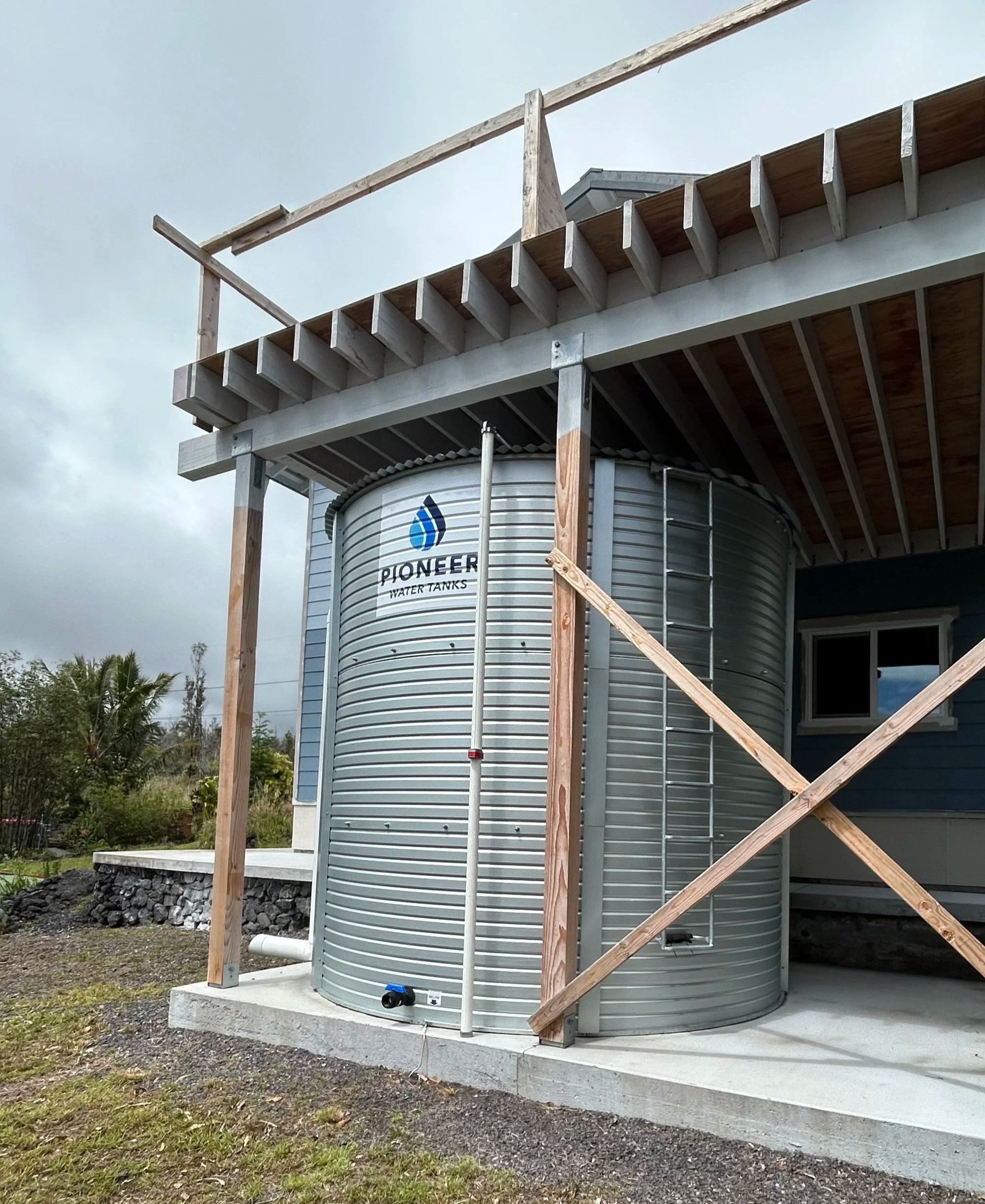 A water tank labeled 'Pioneer Water Tanks' is being installed on a concrete foundation outside a building, with wooden framing supporting a deck or balcony above.