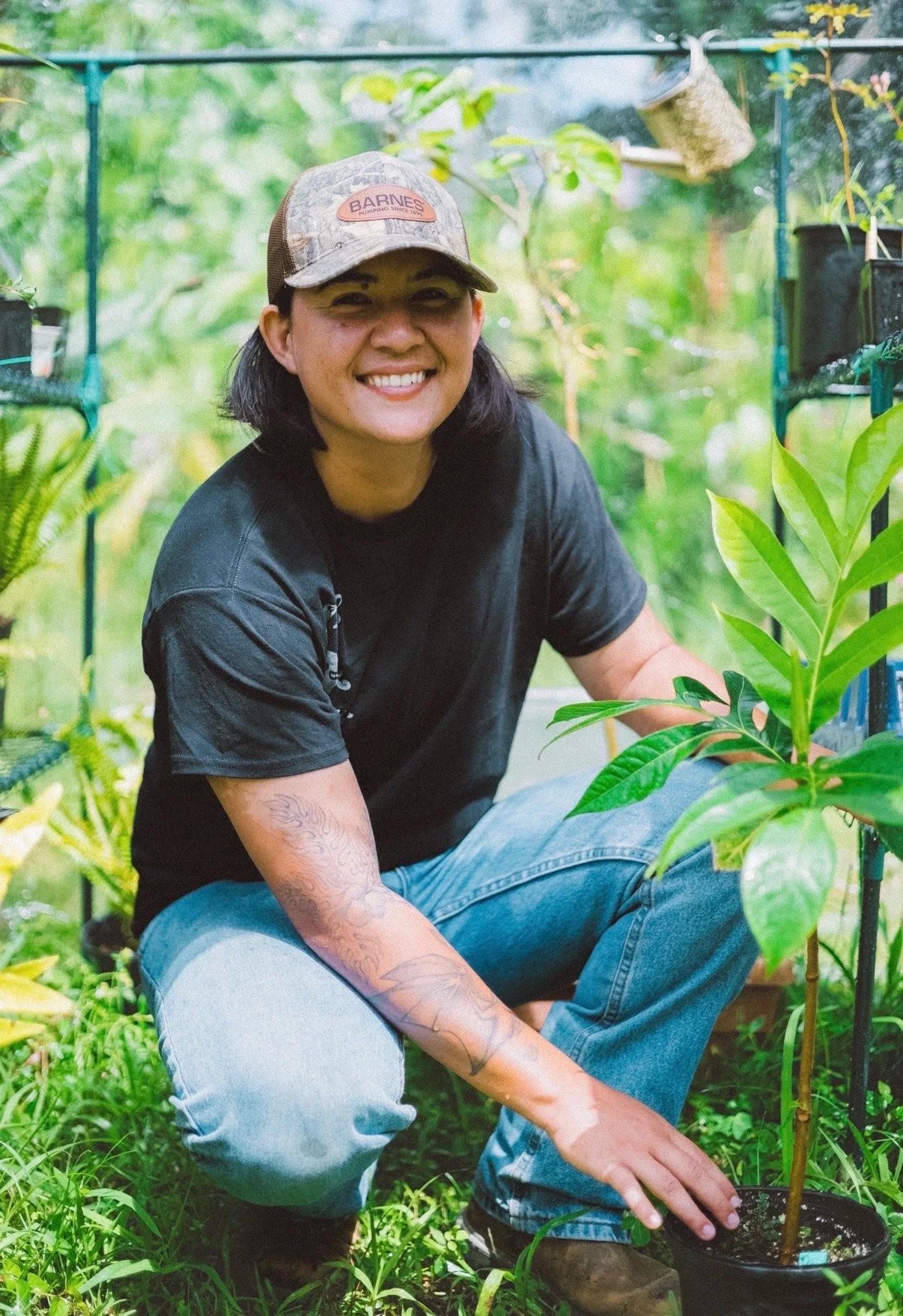 A woman kneeling in a greenhouse, smiling at the camera while tending to a potted plant with broad green leaves. She is wearing a camouflage cap, black T-shirt, and blue jeans.