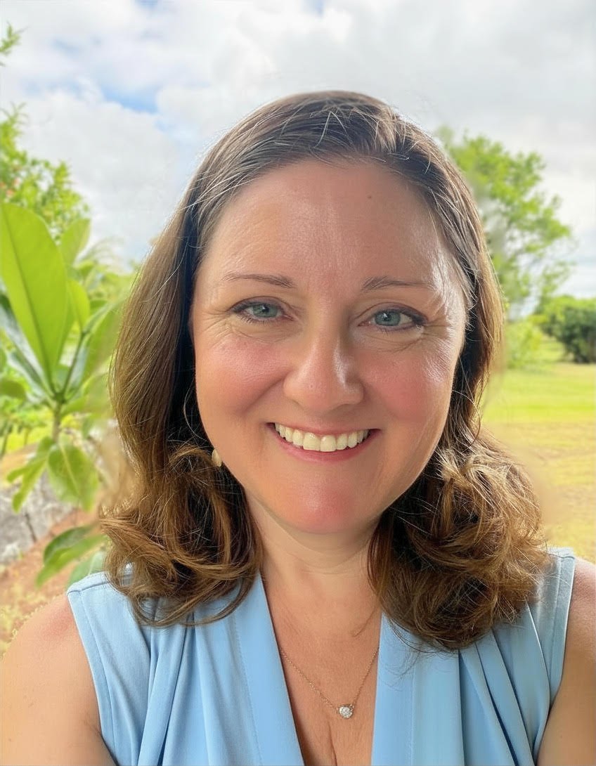 A woman with shoulder-length brown hair and blue eyes smiling outdoors, wearing a light blue sleeveless top, with greenery and a cloudy sky in the background.