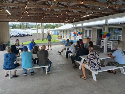 People gathered under a covered outdoor pavilion, sitting on white benches, attending a small outdoor event or party decorated with balloons, with a building, cars, and a grassy area in the background.