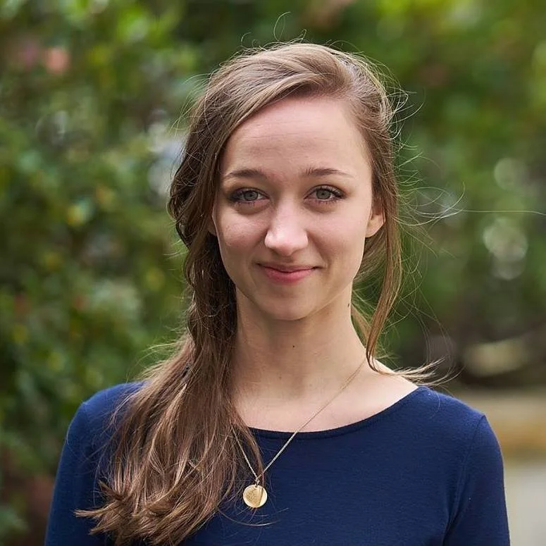 A young woman with long light brown hair, wearing a navy blue top and a gold pendant necklace, standing outdoors with green foliage in the background.