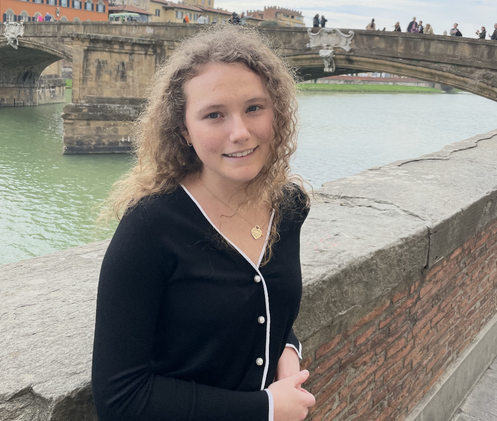 Young woman with curly hair smiling near a stone bridge over a river with colorful buildings in the background.