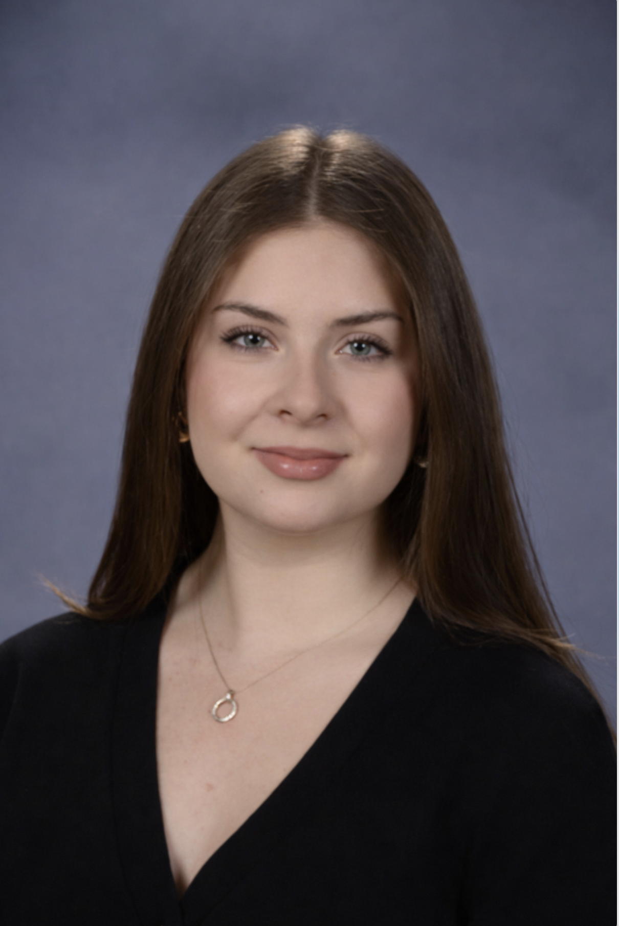 A young woman with long brown hair, blue eyes, and fair skin is smiling slightly at the camera. She is wearing a black top and a delicate necklace with a circular pendant. The background is a gradient of dark blue to gray.