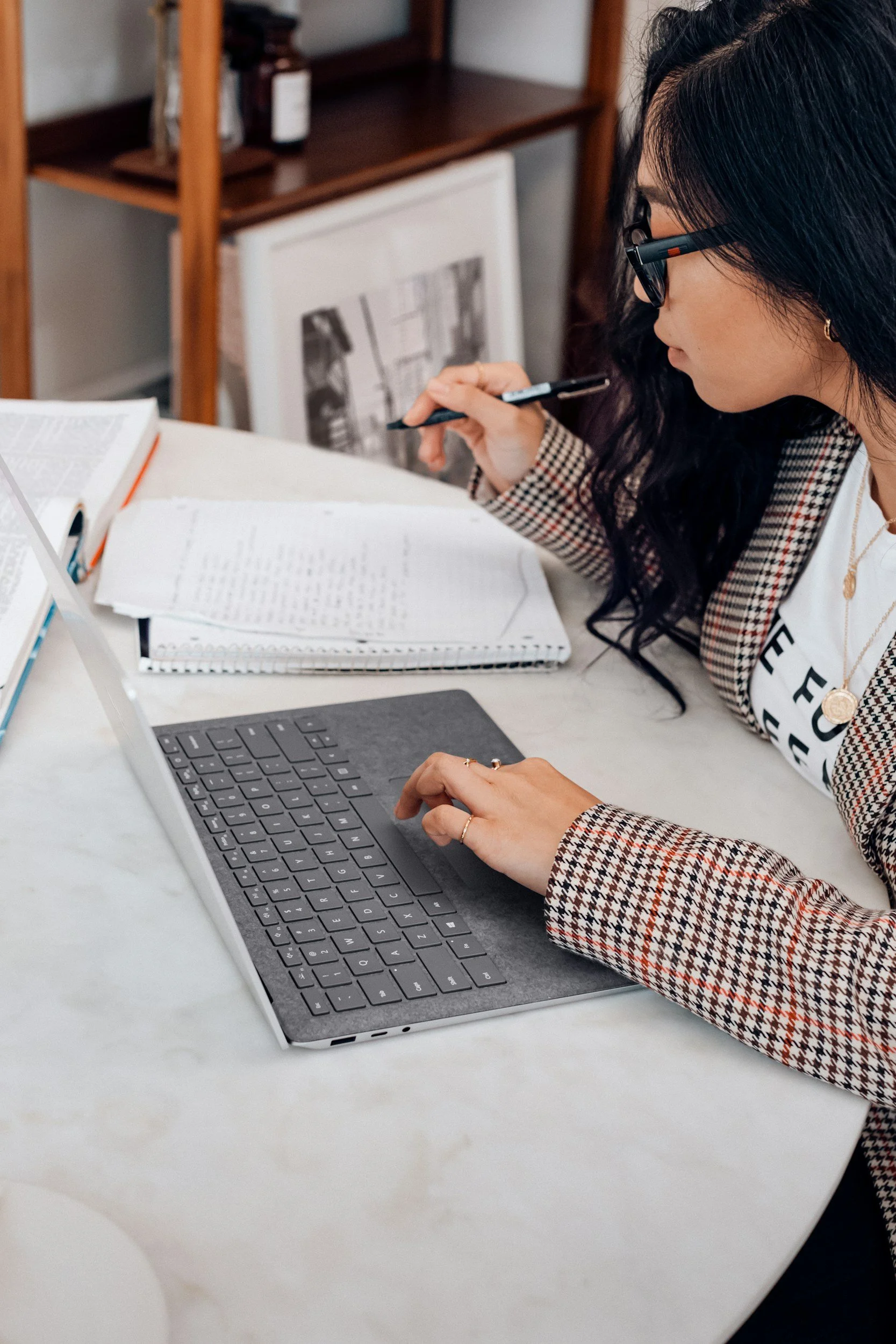 A woman with black hair and glasses working on a laptop at a white table, surrounded by notebooks, papers, and books, with a wooden shelf in the background.