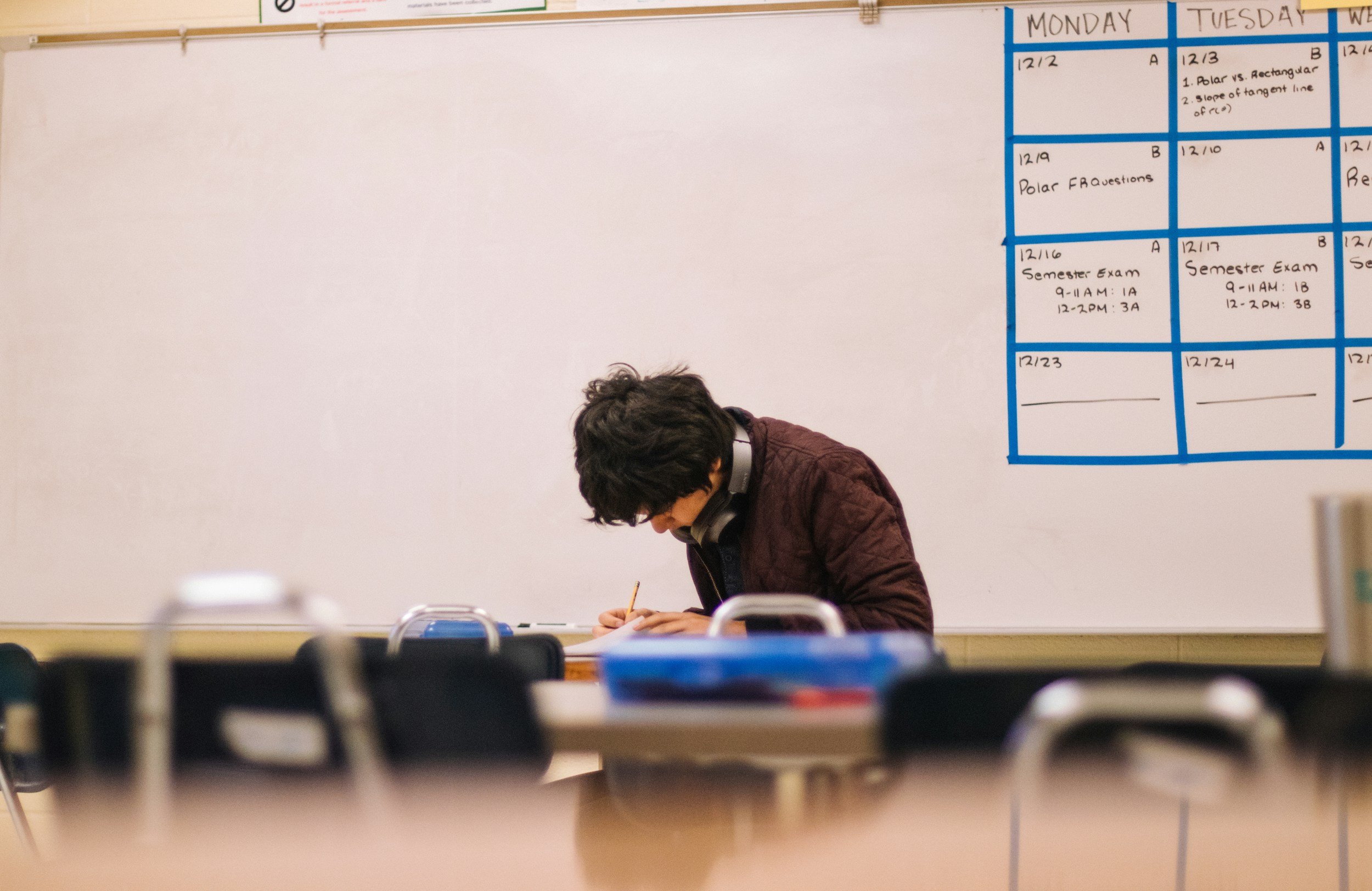 A student with curly hair wearing headphones, sitting at a desk and writing in a notebook in a classroom with a whiteboard and a schedule on the wall.