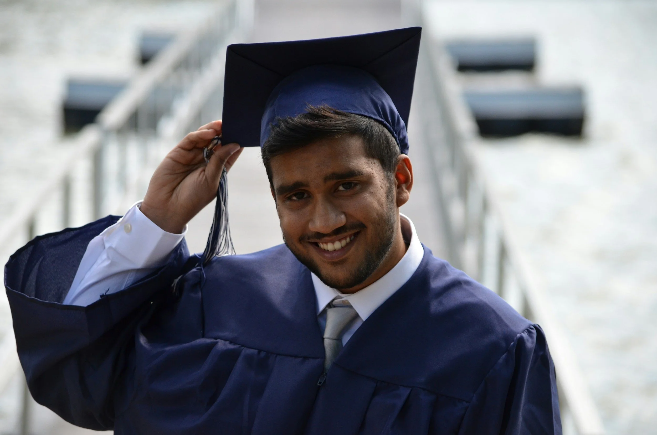 A young man in a navy graduation gown and cap, smiling, adjusting his cap with his right hand.