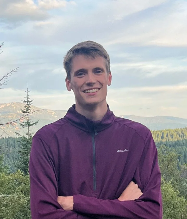 A young man smiling outdoors with mountains and trees in the background.