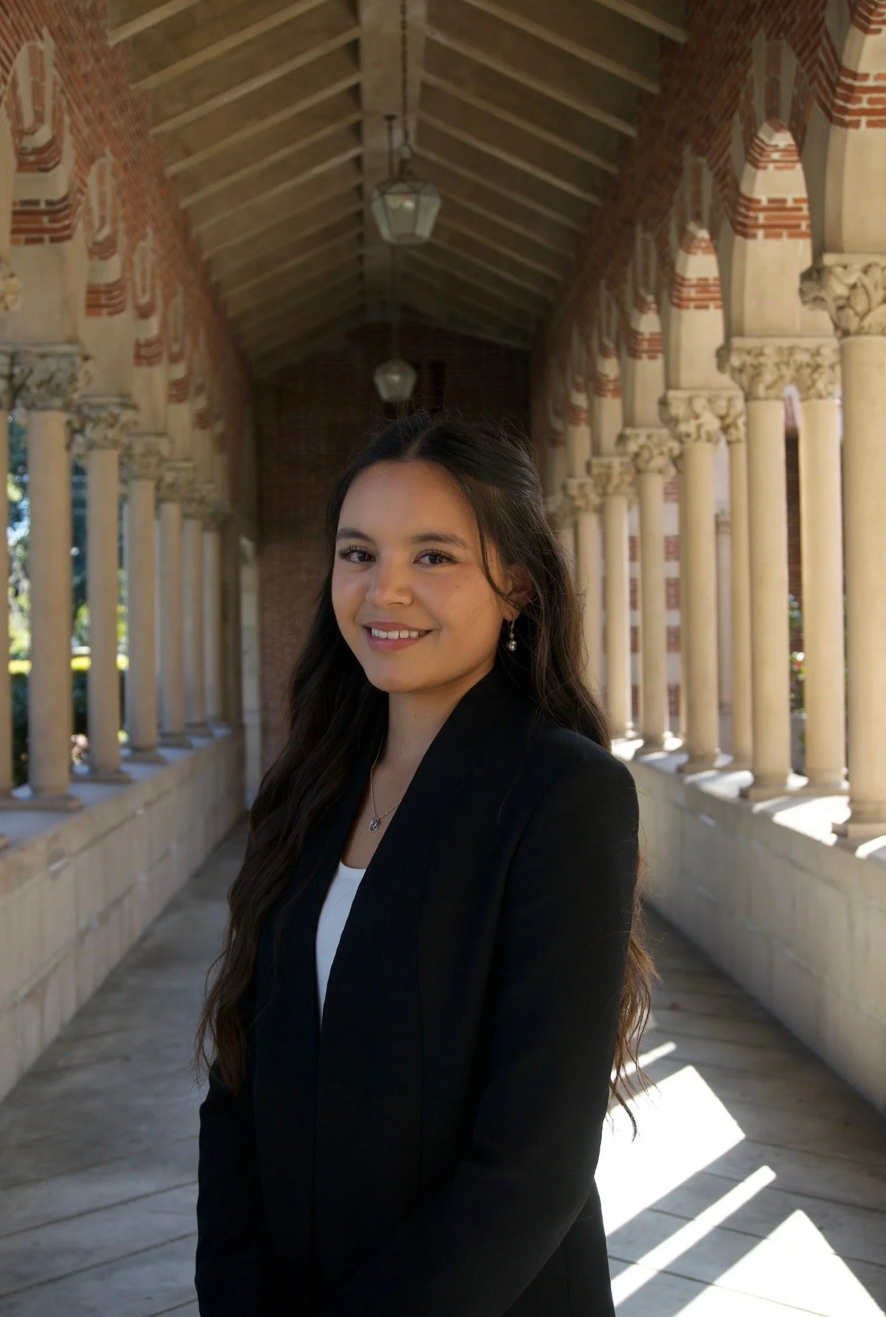 A young woman with long wavy brown hair stands in a covered outdoor walkway lined with columns, wearing a black blazer and smiling at the camera.