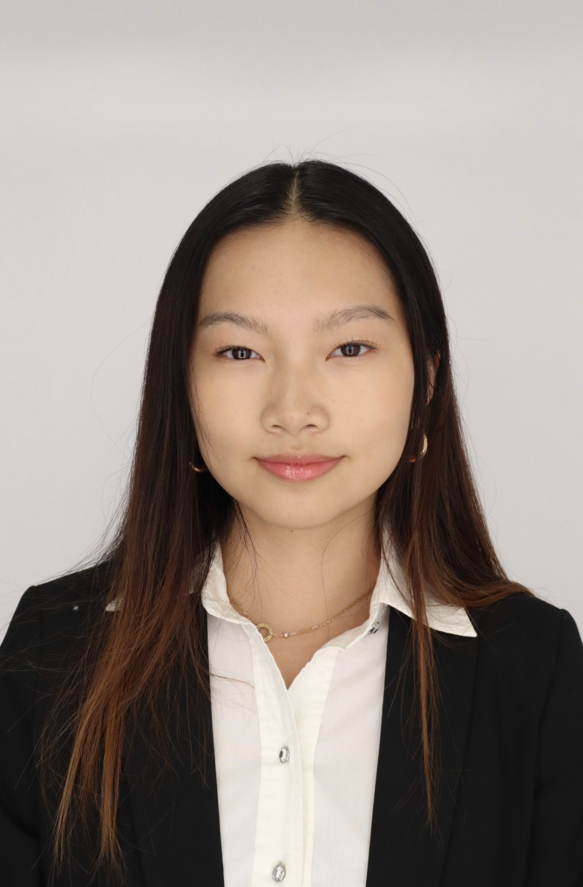 A young woman with long dark hair, wearing a white shirt and black blazer, smiling in front of a plain white background.
