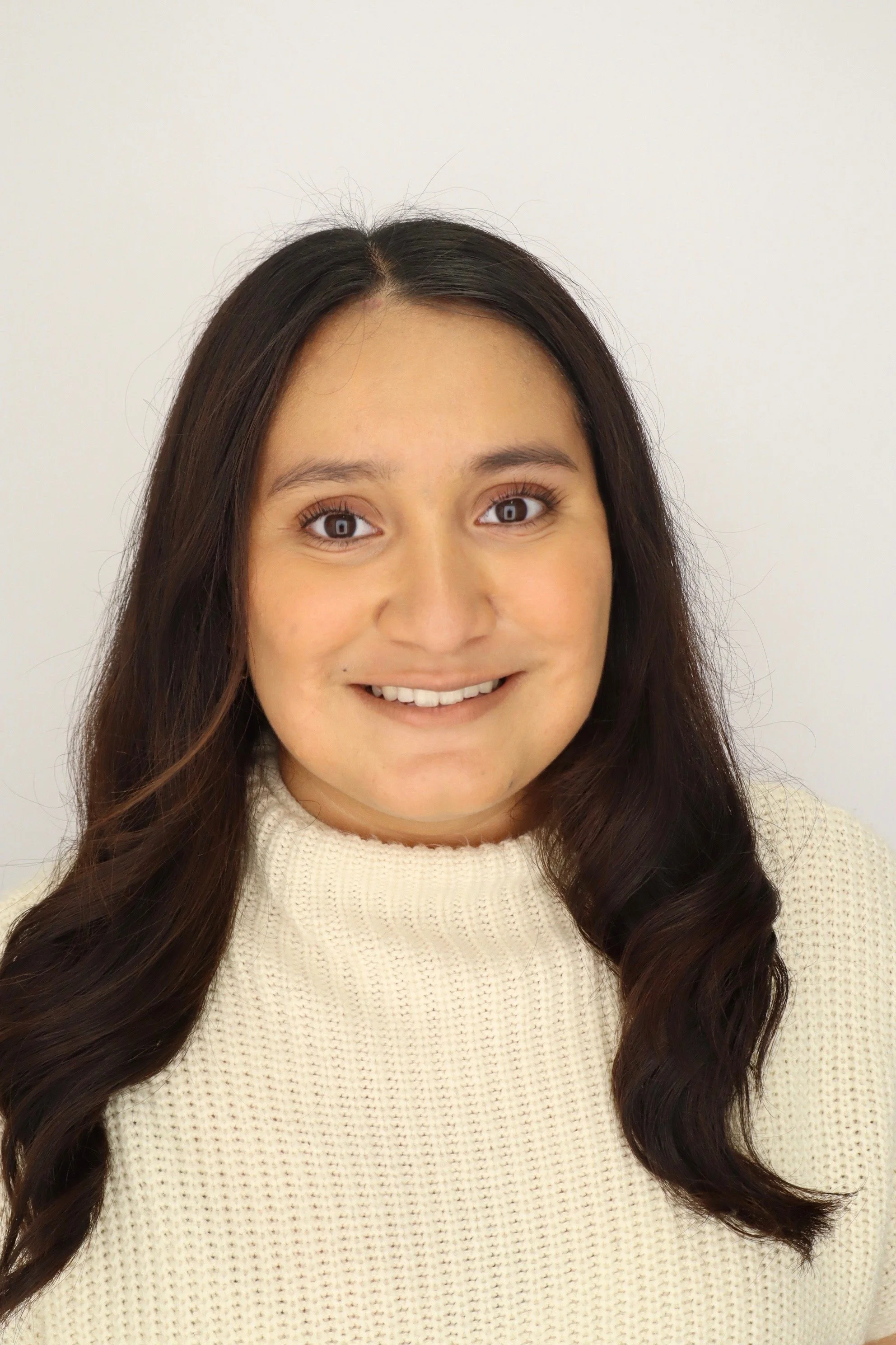 Smiling woman with long dark hair wearing a cream-colored sweater against a light background.